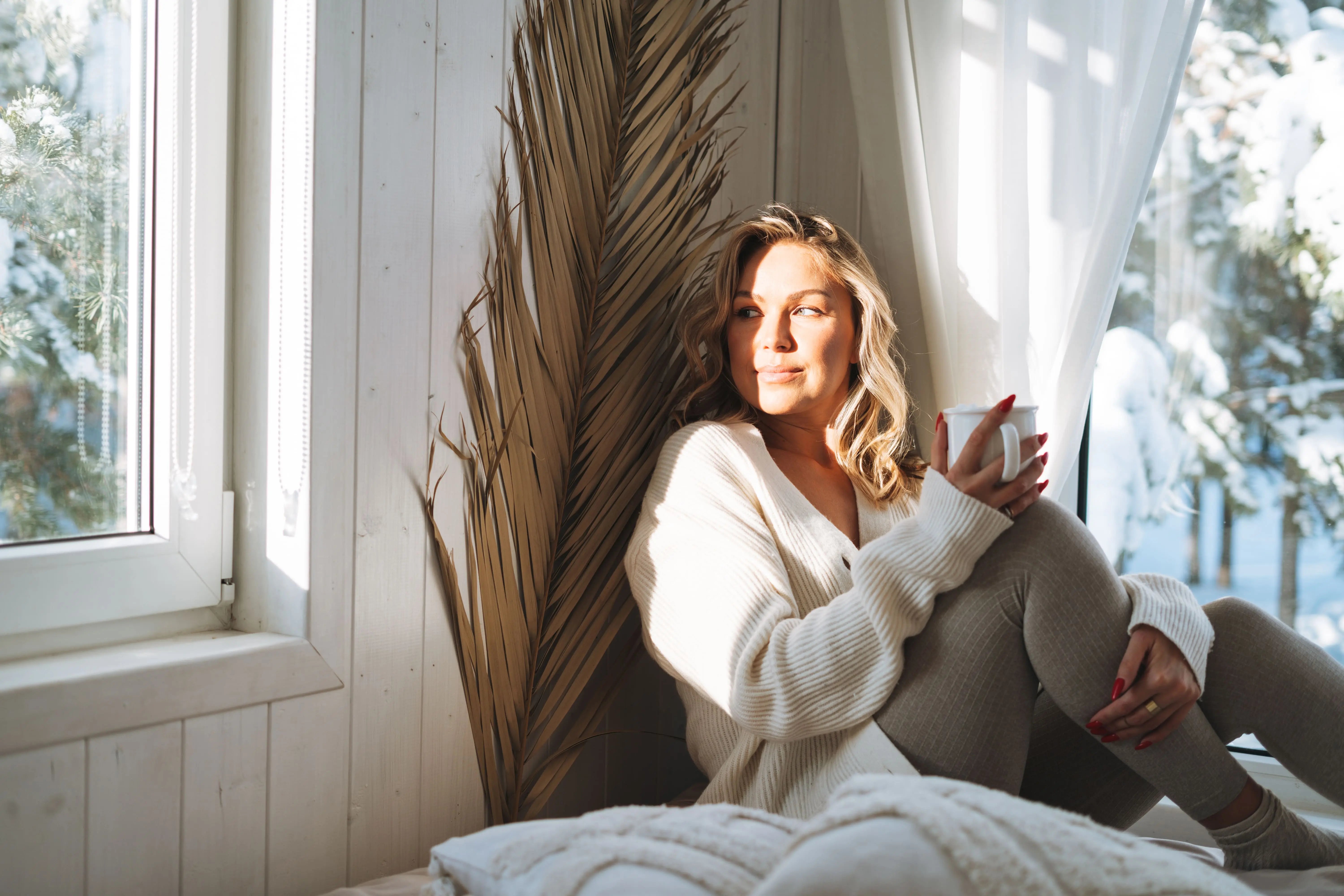 Blonde Frau mit einer Tasse in der Hand sitz auf Fenster und wirkt entspannt.